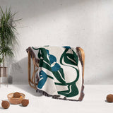 Decorative Matisse style blue floral cotton woven throw blanket with green and blue abstract design draped over a chair against a light gray wall.