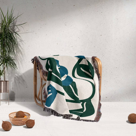 Decorative Matisse style blue floral cotton woven throw blanket with green and blue abstract design draped over a chair against a light gray wall.