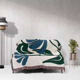Decorative Matisse style blue floral cotton woven throw blanket with leaf pattern draped over a chair against a white wall.