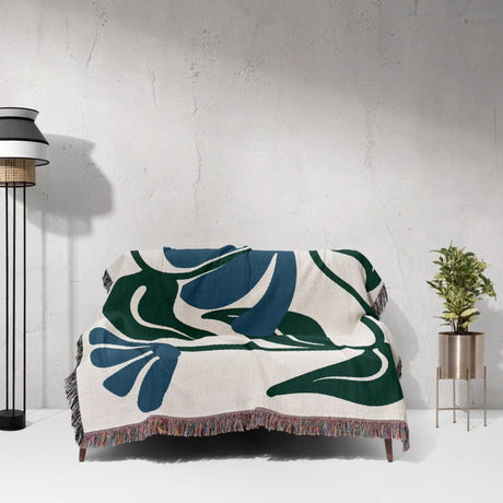 Decorative Matisse style blue floral cotton woven throw blanket with leaf pattern draped over a chair against a white wall.
