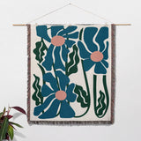 Floral patterned Matisse style blue flowers cotton woven throw blanket with blue flowers and green leaves on a white wall.