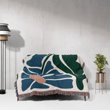 Floral-patterned Matisse style floral cotton woven throw blanket for couch draped over a couch against a white wall with a lamp and plant in the background.