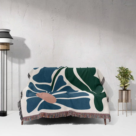 Floral-patterned Matisse style floral cotton woven throw blanket for couch draped over a couch against a white wall with a lamp and plant in the background.