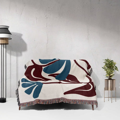 Decorative Matisse style red and blue cotton woven throw blanket with abstract design on a white surface, with a lamp and plant in the background.