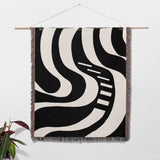 Black and white modern abstract art woven blanket throw tapestry hanging on a wall with a plant in the corner.