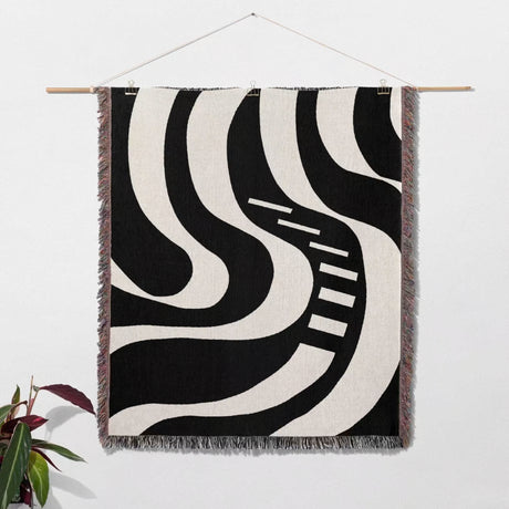 Black and white modern abstract art woven blanket throw tapestry hanging on a wall with a plant in the corner.