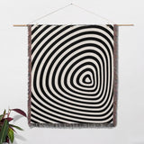 Black and white optical illusion hole woven blanket throw tapestry hanging on a white wall with a plant in the corner.