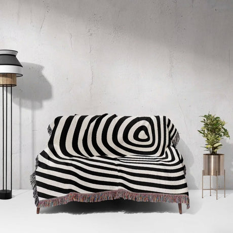 Black and white optical illusion hole woven blanket throw tapestry draped over a couch against a white wall.
