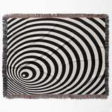 Black and white optical illusion wormhole woven blanket throw tapestry with fringed edges flat lay on a white background.