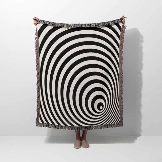 Black and white optical illusion wormhole woven blanket throw tapestry draped over a person on a white background.