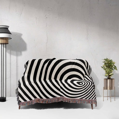 Black and white optical illusion wormhole woven blanket throw tapestry draped over a sofa against a white wall.