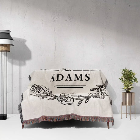 Personalized 2 year anniversary gift - Mr and Mrs woven throw blanket with 'ADAMS' and floral design on a white surface with a lamp and plant in the background.
