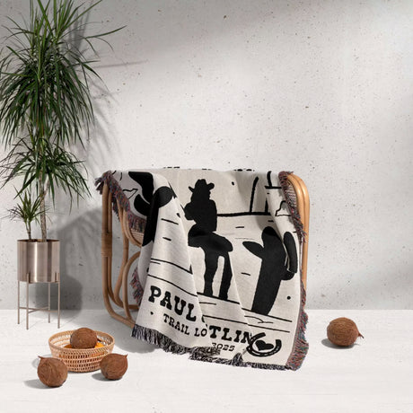 Personalized cowboy and cowgirl cotton anniversary gift woven throw blanket with western design draped over a chair, with a plant and coconuts in the background.