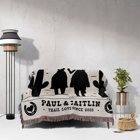 Personalized cowboy and cowgirl cotton anniversary gift woven throw blanket with black silhouettes and text draped over a couch in a room with a lamp and plant.