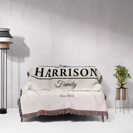 Personalized family name and address blanket with 'Harrison Family' text draped over a sofa against a white wall.