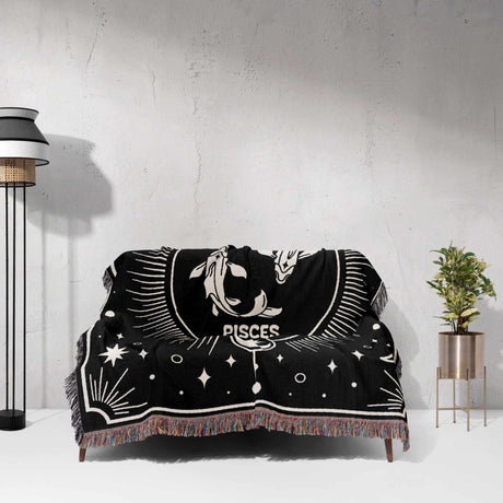 The Pisces Zodiac Blanket is displayed on a couch, showcasing its full design. The intricate black and white pattern features the Pisces symbol and celestial elements, providing a decorative and cozy look.