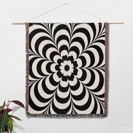 Black and white psychedelic flower woven blanket throw tapestry hanged on a white wall with a plant in the corner.