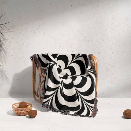 Black and white psychedelic flower woven blanket throw tapestry draped over a chair against a light gray background.