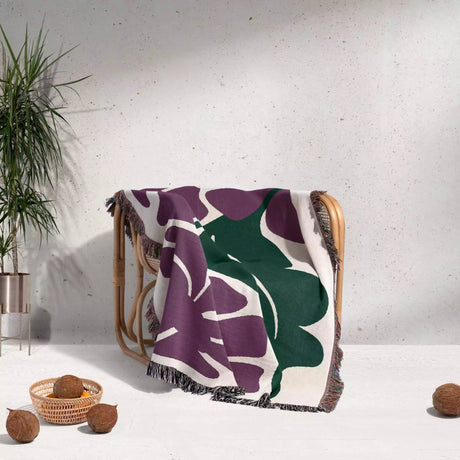 Decorative purple Matisse flowers woven throw blanket with purple and green leaf pattern draped over a chair against a light gray wall.