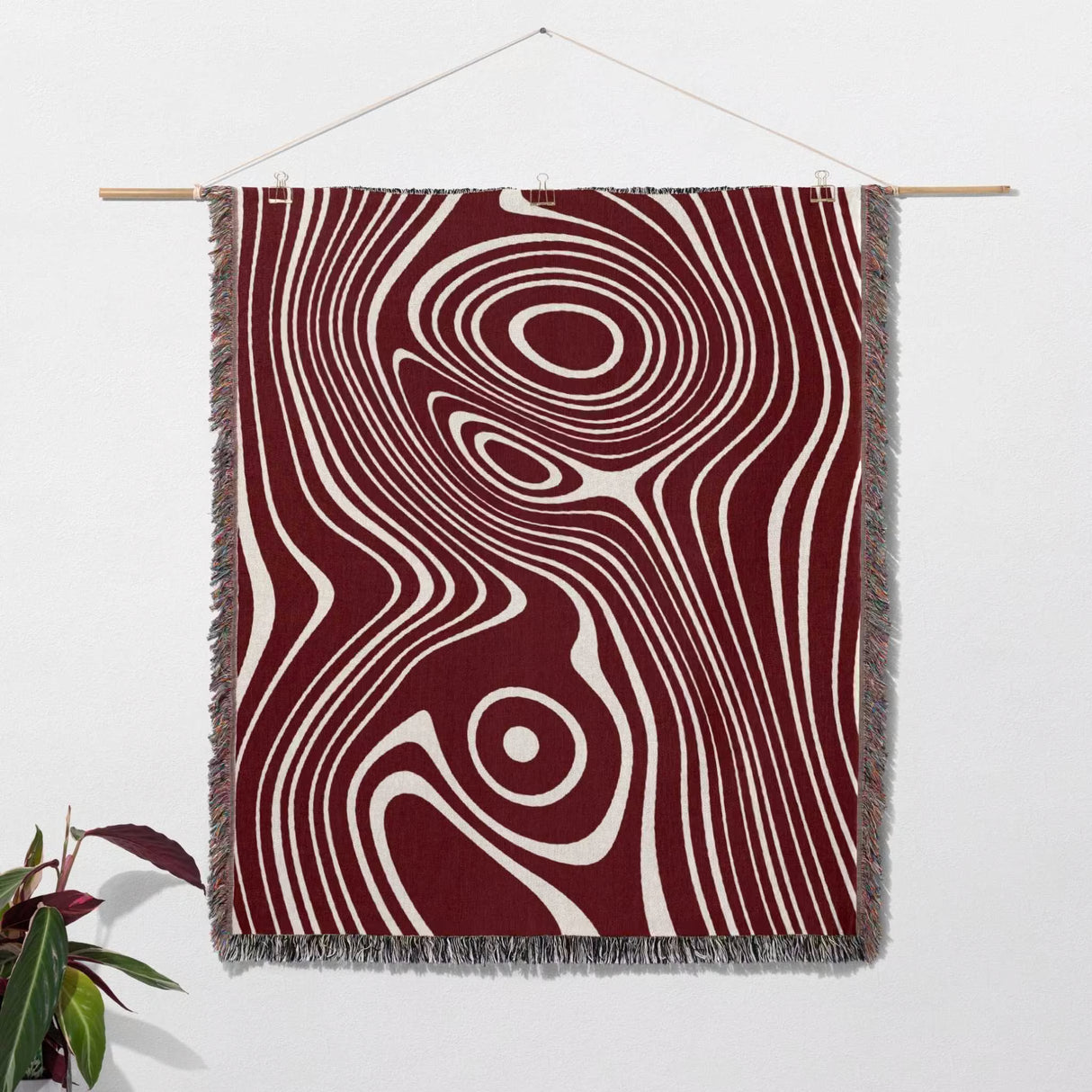 Red abstract woven throw blanket with a swirling pattern in red and white on a wooden rod against a white wall.