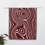 Red abstract woven throw blanket with a swirling pattern in red and white on a wooden rod against a white wall.