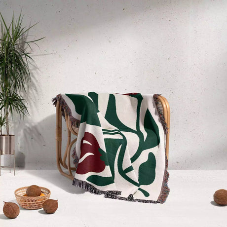 Red and green Matisse flower woven throw blanket decorative blanket with leaf pattern draped over a chair against a light gray wall.