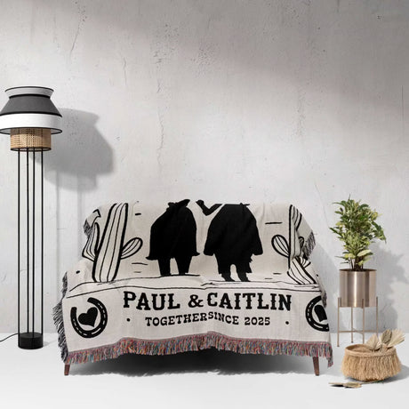 Romantic cowboy and cowgirl cotton anniversary gift – woven throw blanket with silhouettes of a couple and text 'PAUL & CAITLIN TOGETHER SINCE 2023' draped over a couch in a room.