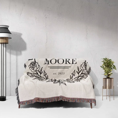 Rustic Mr and Mrs 2nd year anniversary gift woven throw blanket with 'MOORE est. 2023' design on a couch against a gra wall.