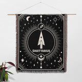 The Sagittarius Zodiac blanket is hung on a wall like a tapestry, showcasing its decorative potential. The black and white woven design stands out, making it a unique piece of wall art with the central arrow symbol and celestial motifs.