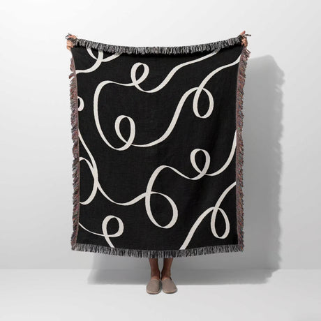 Black and white scribble lined woven blanket throw with fringe held by a person on a white background.