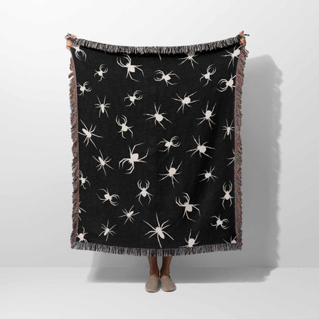 A full view of the Halloween woven blanket being held up vertically. The blanket displays a detailed pattern of white spiders on a black background, with a colorful fringe lining the edges. The dark gothic theme is emphasized by the bold contrast of colors.