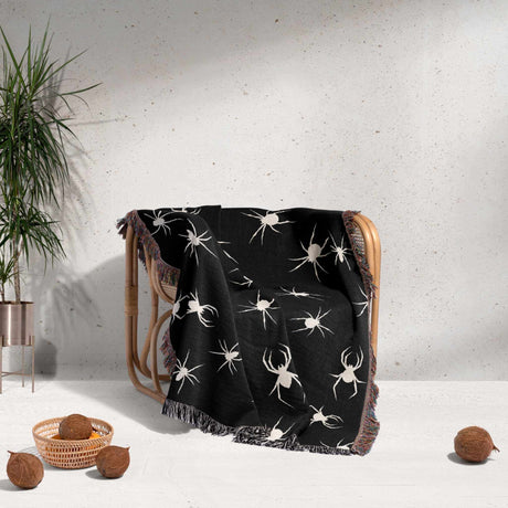 A cozy scene with a Halloween-themed woven blanket draped over a wooden chair. The blanket, featuring a dark black base with white spider motifs, contrasts with the warm, earthy tones of the room. The multicolor fringe is visible at the edges.