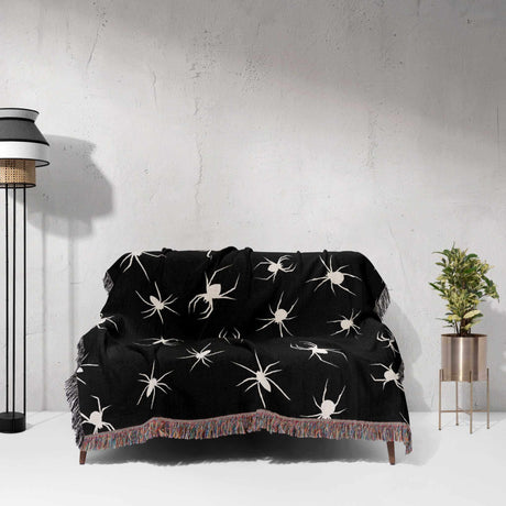 The Halloween woven blanket spread out on a modern couch. The black background and white spider pattern create a striking contrast, making it a perfect dark gothic decor piece. The multicolor fringe adds a unique touch to the overall aesthetic.