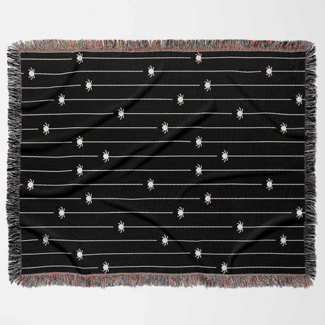 This flat-lay image displays the entire Spooky Halloween Woven Blanket in a horizontal position. The design features multiple off-white spiders hanging from thin lines on a solid black background. The blanket’s multicolored fringe adds a playful touch to the eerie spider pattern, making it a perfect Halloween-themed throw for any space.