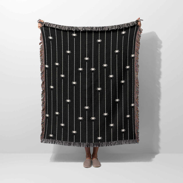 In this image, the Spooky Halloween Woven Blanket is held vertically by a person, providing a sense of its size and design. The blanket, featuring an array of off-white spiders against a black backdrop, is bordered by a colorful fringe. This view highlights the blanket’s spooky yet stylish design, making it clear why it's a must-have for Halloween enthusiasts.