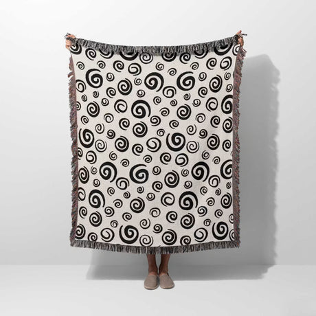 Black and white swirls patterned woven blanket throw with fringe held by a person on a white background.