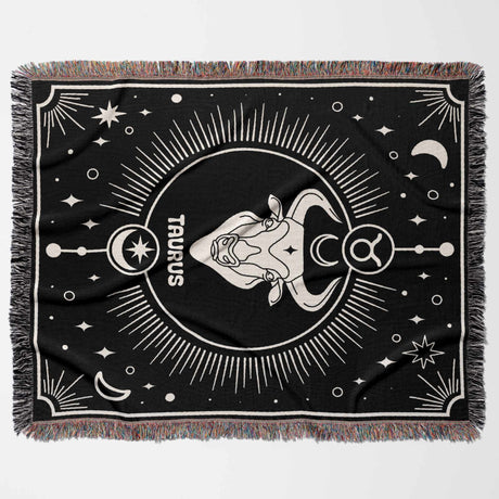 Flat horizontal view of the Taurus Zodiac Blanket displaying the full design. The black and white woven throw blanket features the Taurus zodiac symbol and bull head in the center, surrounded by celestial elements and a multicolor fringe.