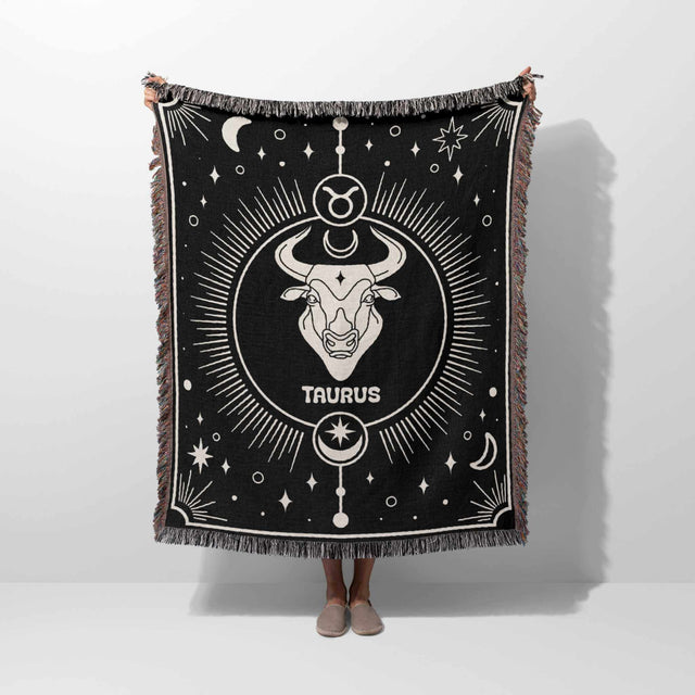 Person holding up the Taurus Zodiac Blanket, displaying its full design. The black and white woven throw blanket with the Taurus sign and celestial elements is shown in full view, highlighting its intricate details and multicolor fringe.