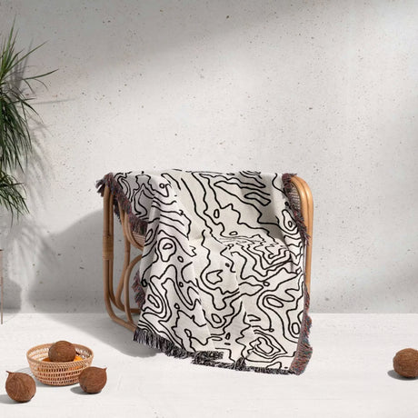 Wooden chair with a black and white topographic map design woven throw blanket draped over it on a light background.