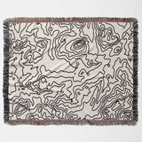 Black and white topographic map design woven throw blanket with black line art pattern flat lay on a light background.