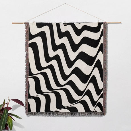 Black and white trendy abstract waves woven blanket throw tapestry with fringe hanging on a wooden rod against a white wall.