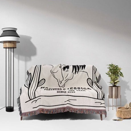 Two horses woven throw blanket cotton anniversary gift with horse design and text draped over a couch in a room with a lamp and plant.