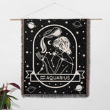 This image shows a Zodiac Sign Blanket used as wall decor, depicting the Aquarius astrological sign. The woven tapestry features a black-and-white illustration with celestial motifs, ideal for adding an astrological touch to your home as both a decorative piece and a cozy blanket.